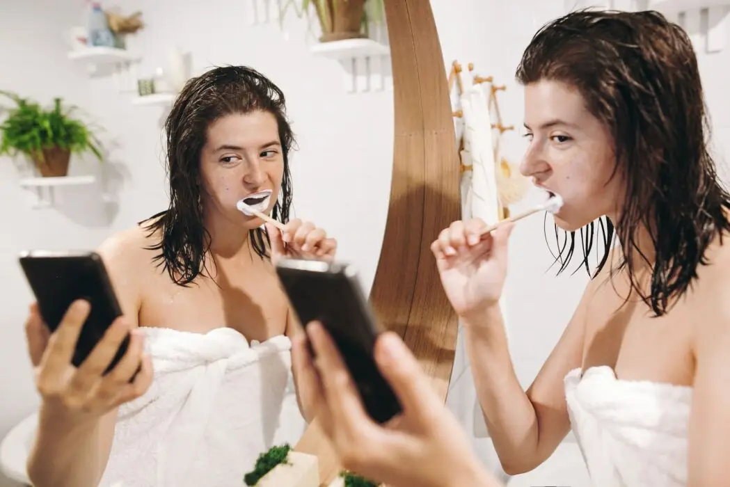 Young happy woman in white towel brushing teeth and looking at smartphone